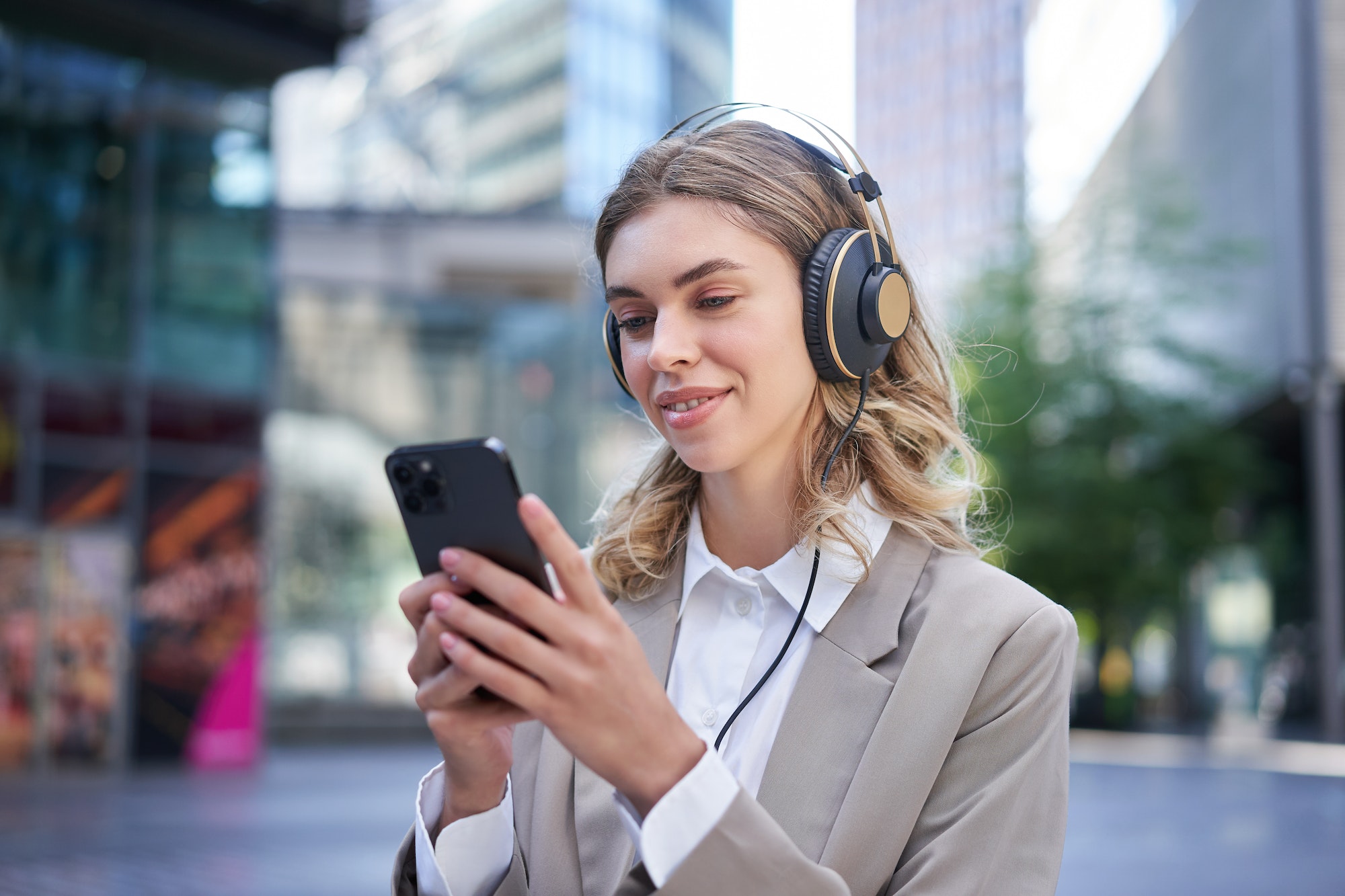 Portrait of smiling young businesswoman listening music in headphones and using mobile phone while