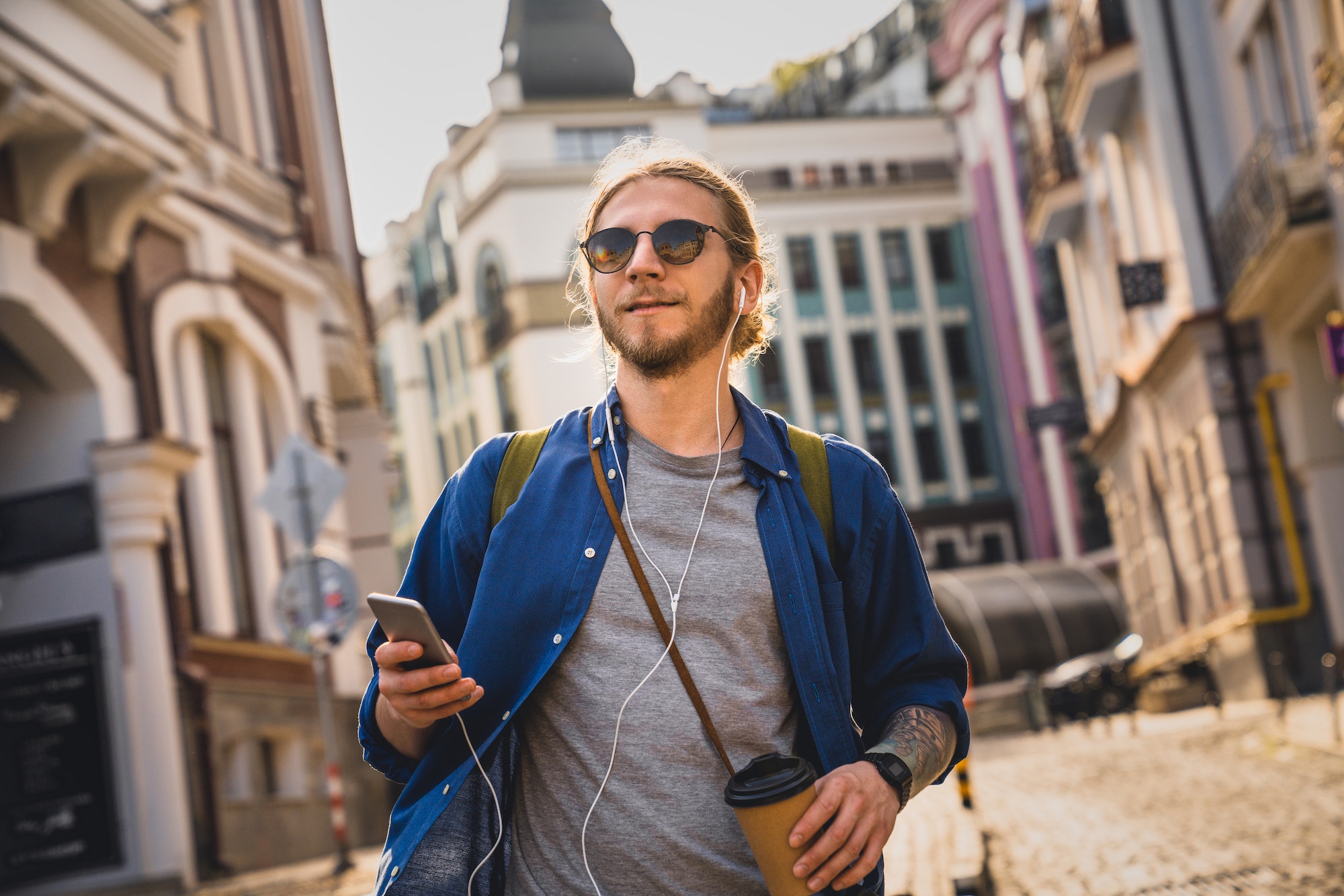 Young beard handsome man drinking coffee and listening music in headphones while walking
