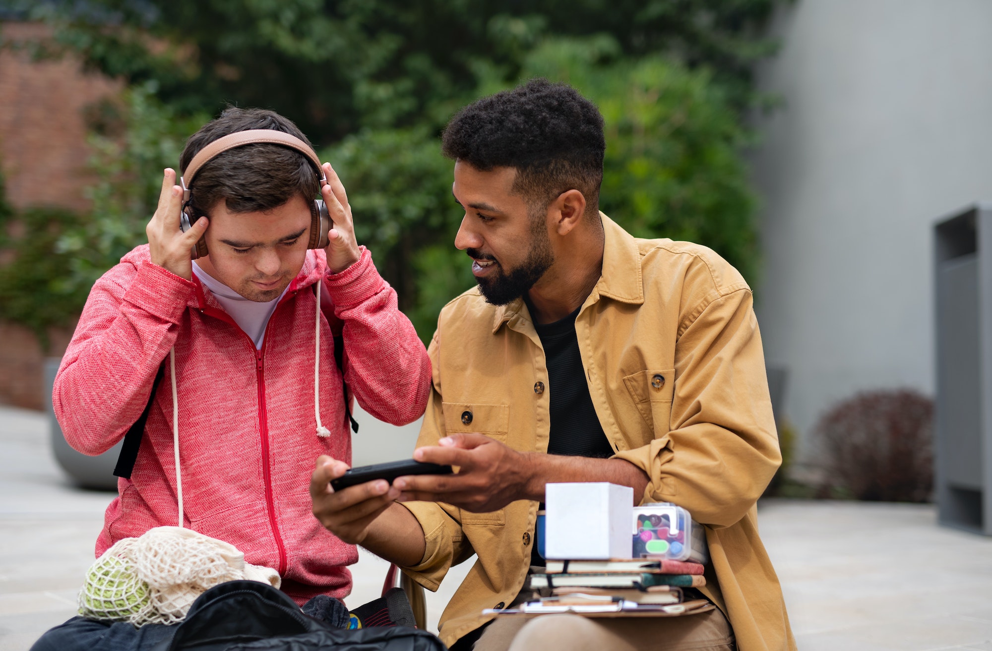 Young man with Down syndrome with his mentoring friend sitting outdoors and listening to music